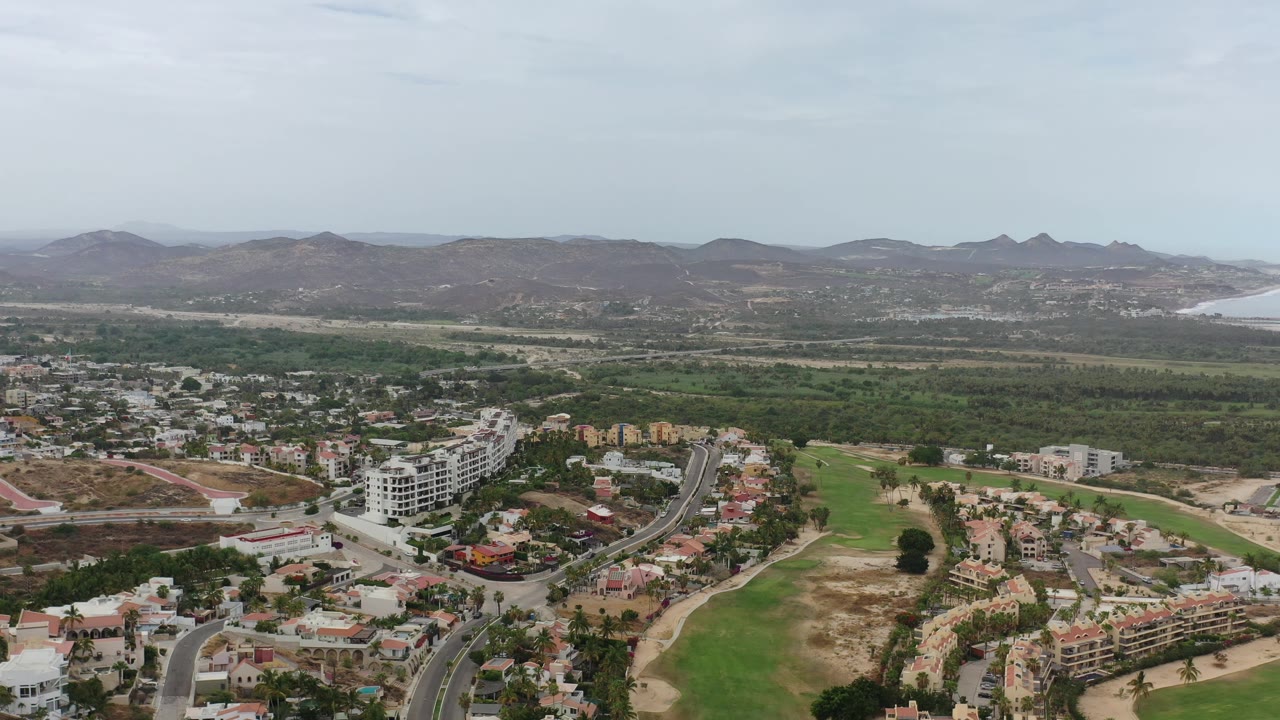 Los Cabos aerial view one second in, centered on a white mid-rise building cluster beside roads, green open land, tree cover, distant brown hills, and no visible people.
