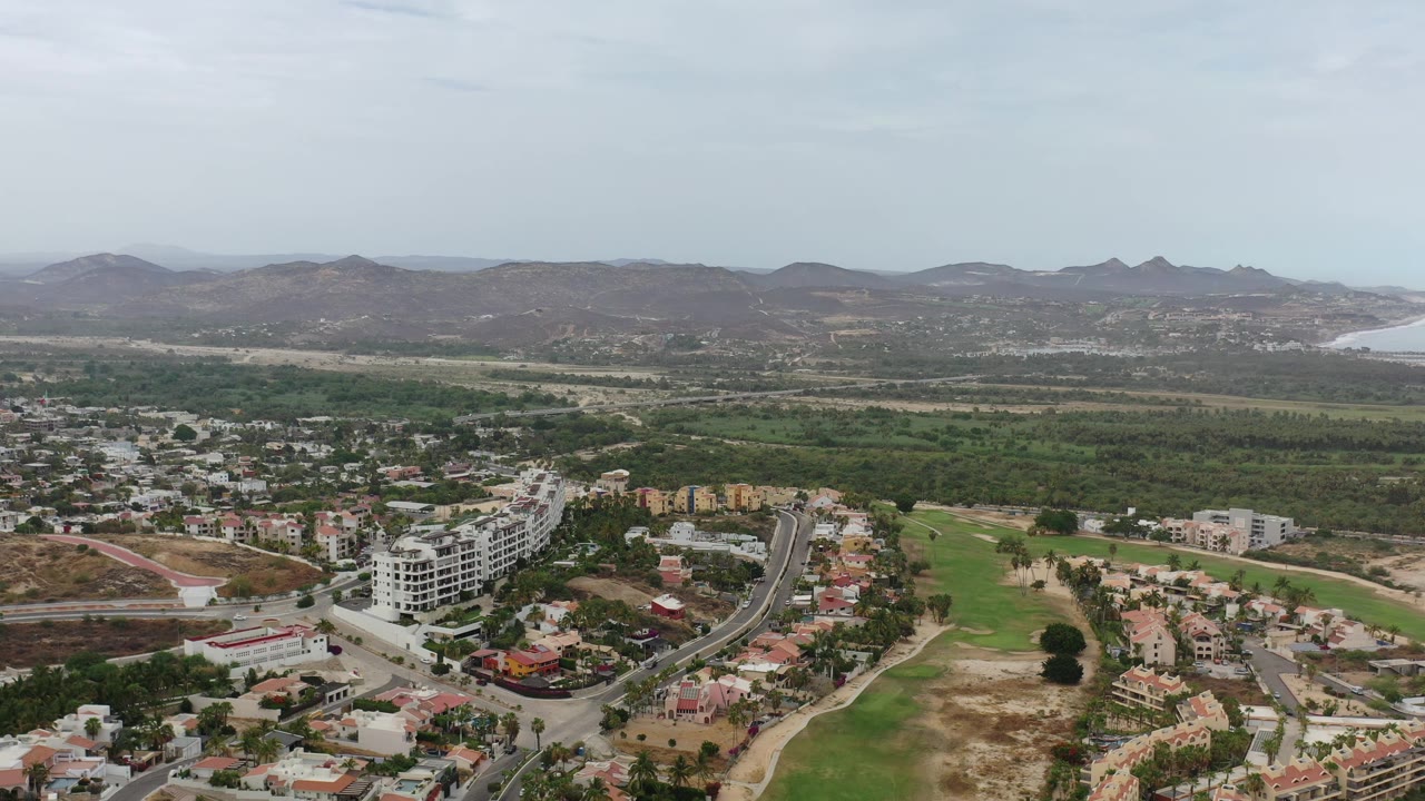 Second aerial frame in Los Cabos, Mexico, showing a similar wide view with a larger central road cluster, apartment-like white buildings, dry lots, hills, bay water, and no visible people.