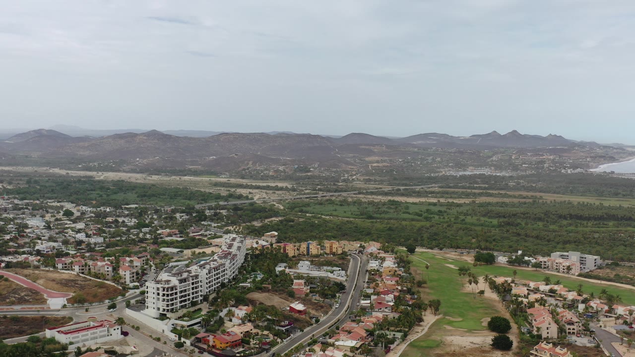 Opening aerial frame over Los Cabos, Mexico, with low-rise buildings and roads in the foreground, green terrain across the middle, distant hills and water, and no visible people.