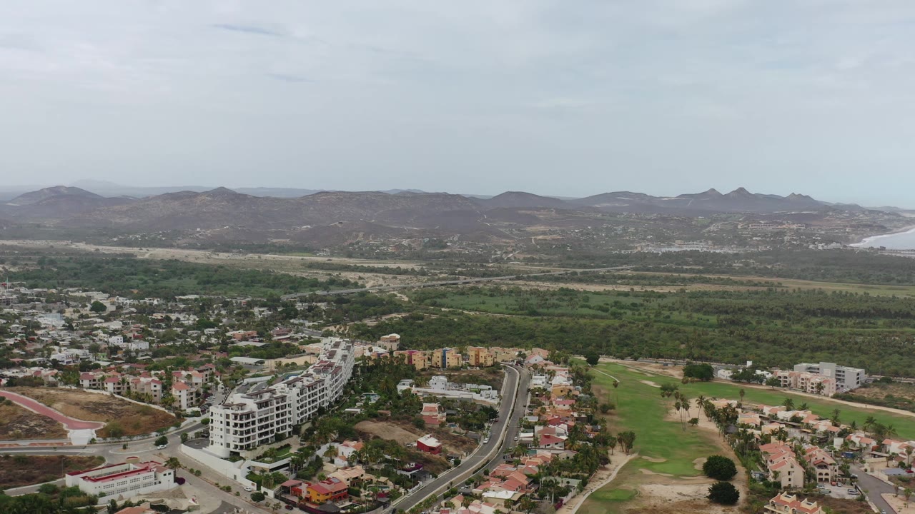 Aerial frame in Los Cabos, Mexico, showing a curved road and clustered rooftops below, broad green vegetation beyond, hazy mountains, distant water, and no visible people.