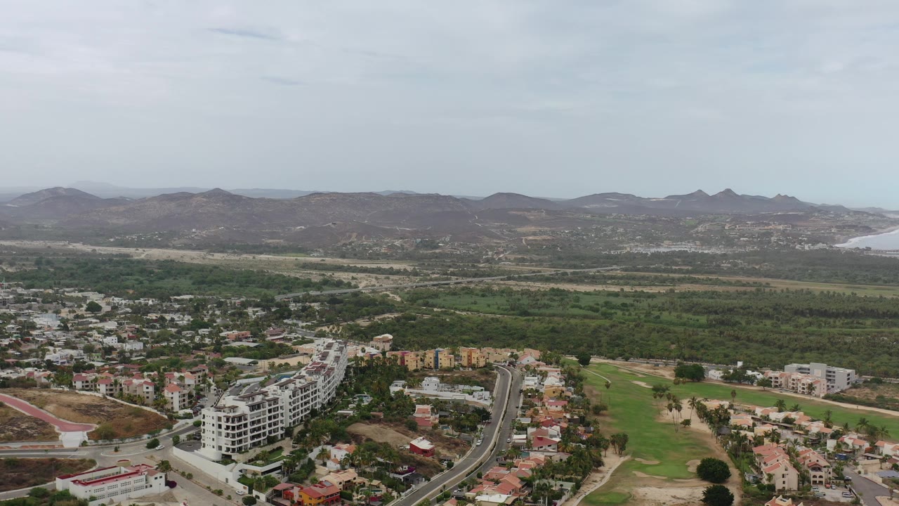 Wide aerial over buildings and green landscape with hills and coastline in the distance.