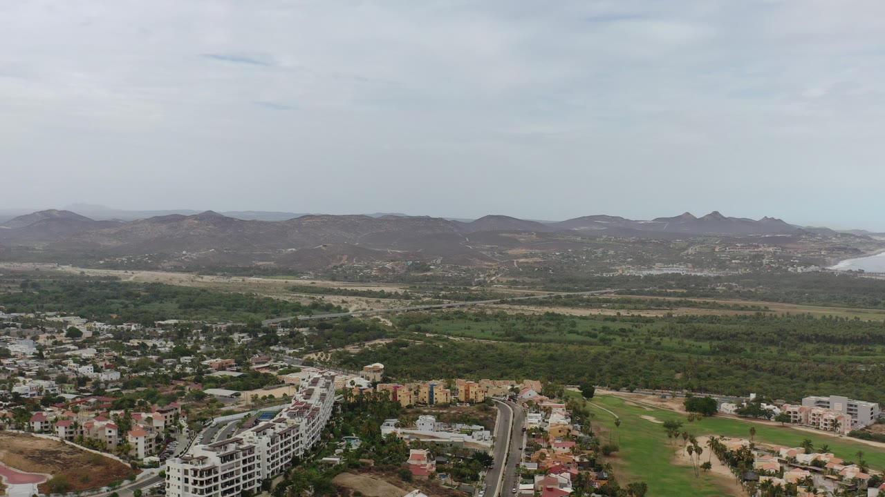Aerial view over low-rise buildings, green land, hills, and distant water.