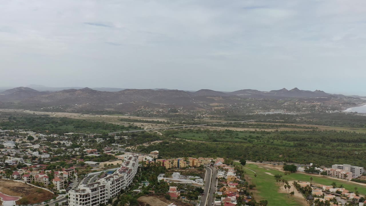 Aerial Los Cabos frame with a denser cluster of buildings at the bottom, green fairway-like land and trees in the midground, and hazy hills near distant water, no visible people.