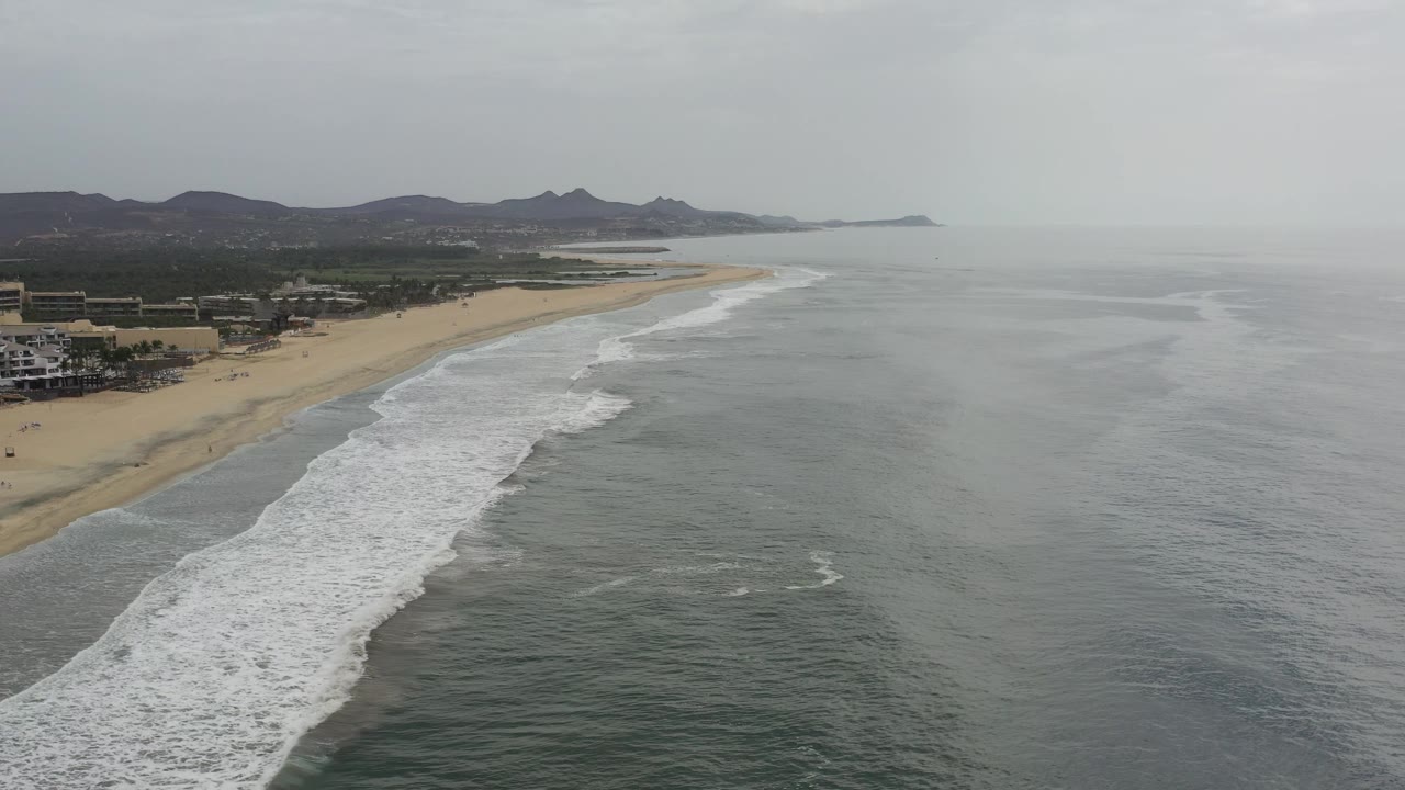 A wide aerial view in Los Cabos, Mexico shows white surf running along a sandy beach, gray ocean water to the right, low buildings at the far left, distant mountains, and no visible people.