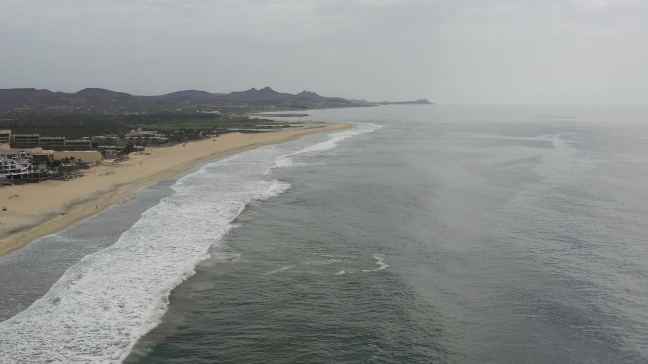 A high view over the Los Cabos shoreline shows broken whitewater edging the sand, open gray-green sea on the right, dark hills in the distance, and no visible people.