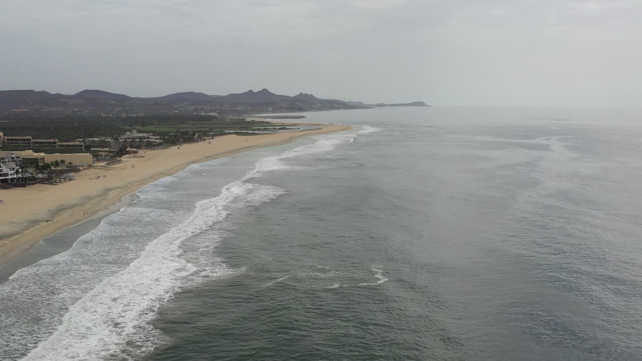Aerial view of waves rolling toward a sandy beach under a gray sky.