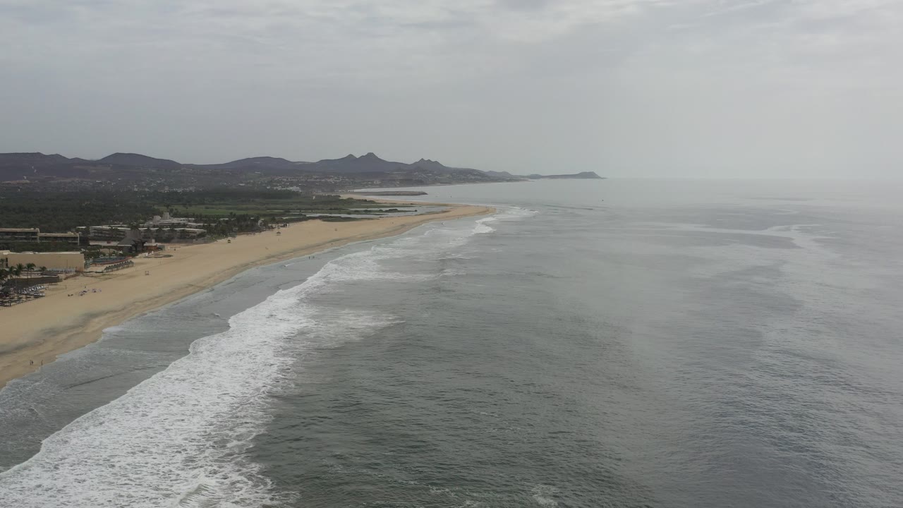 Aerial view of a sandy beach, surf, ocean, distant hills, and gray sky.