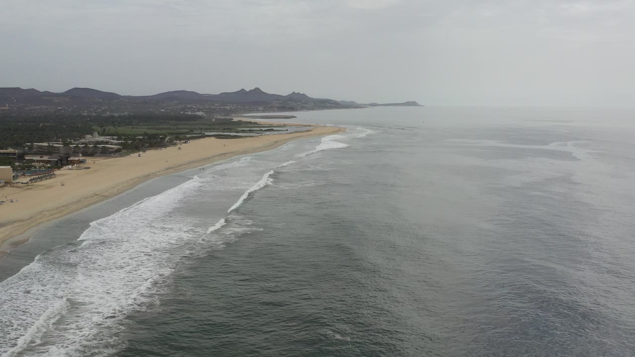 Aerial view of surf reaching a wide beach beside gray ocean.