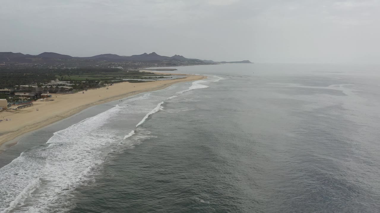 Aerial Los Cabos coastline view with white foam bands curving toward the sand, beachfront greenery and low structures inland, hazy mountains beyond, and no visible people.