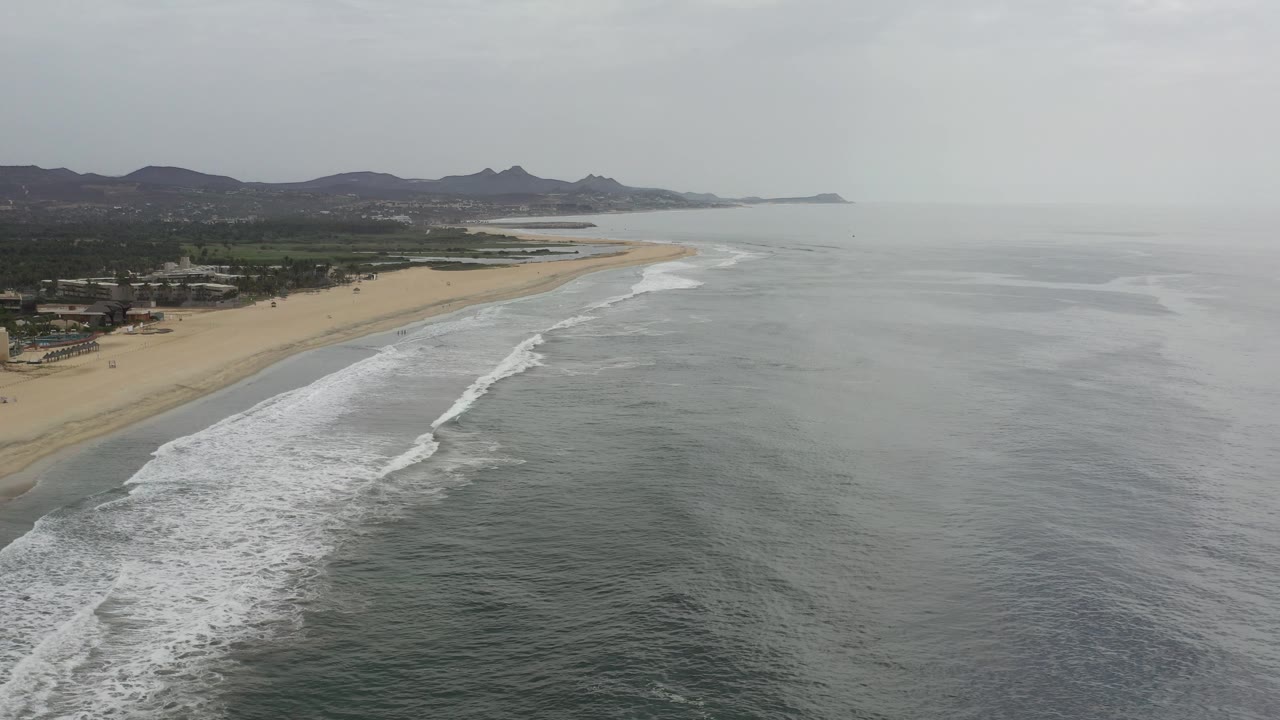 Aerial coastline with ocean, beach, palms, and distant hills.