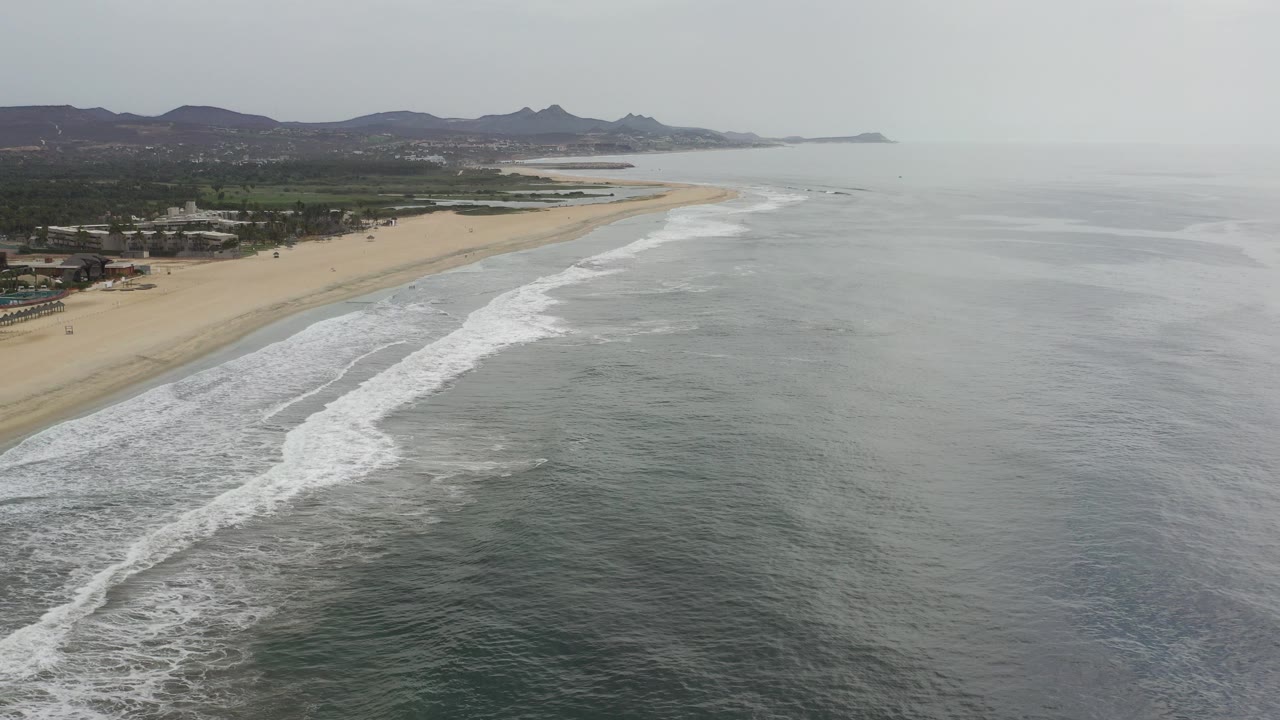 Aerial view of surf beside a sandy beach and distant mountains.