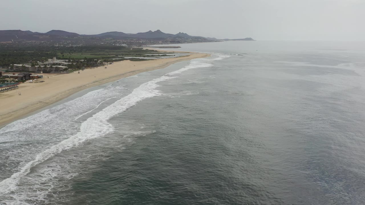 Aerial view along the Los Cabos shoreline as a long band of white surf curves beside the sand, with green land and low mountains under hazy daylight and no visible people.
