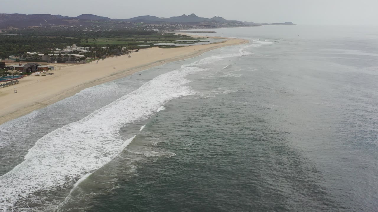 Opening aerial frame over the Los Cabos shoreline, with white surf sweeping diagonally across green-gray ocean toward a long sandy beach and no visible people.