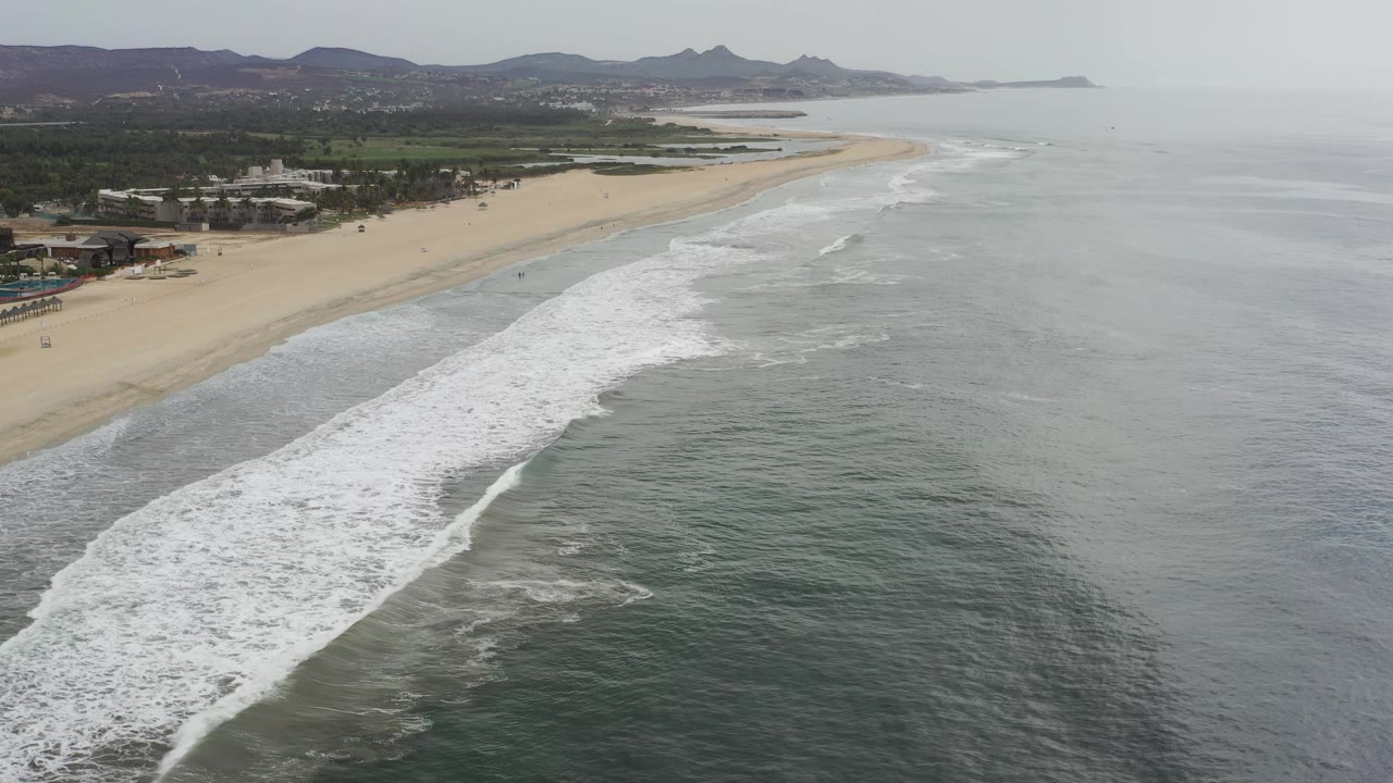 Drone-like view of a beach with white surf and distant hills.