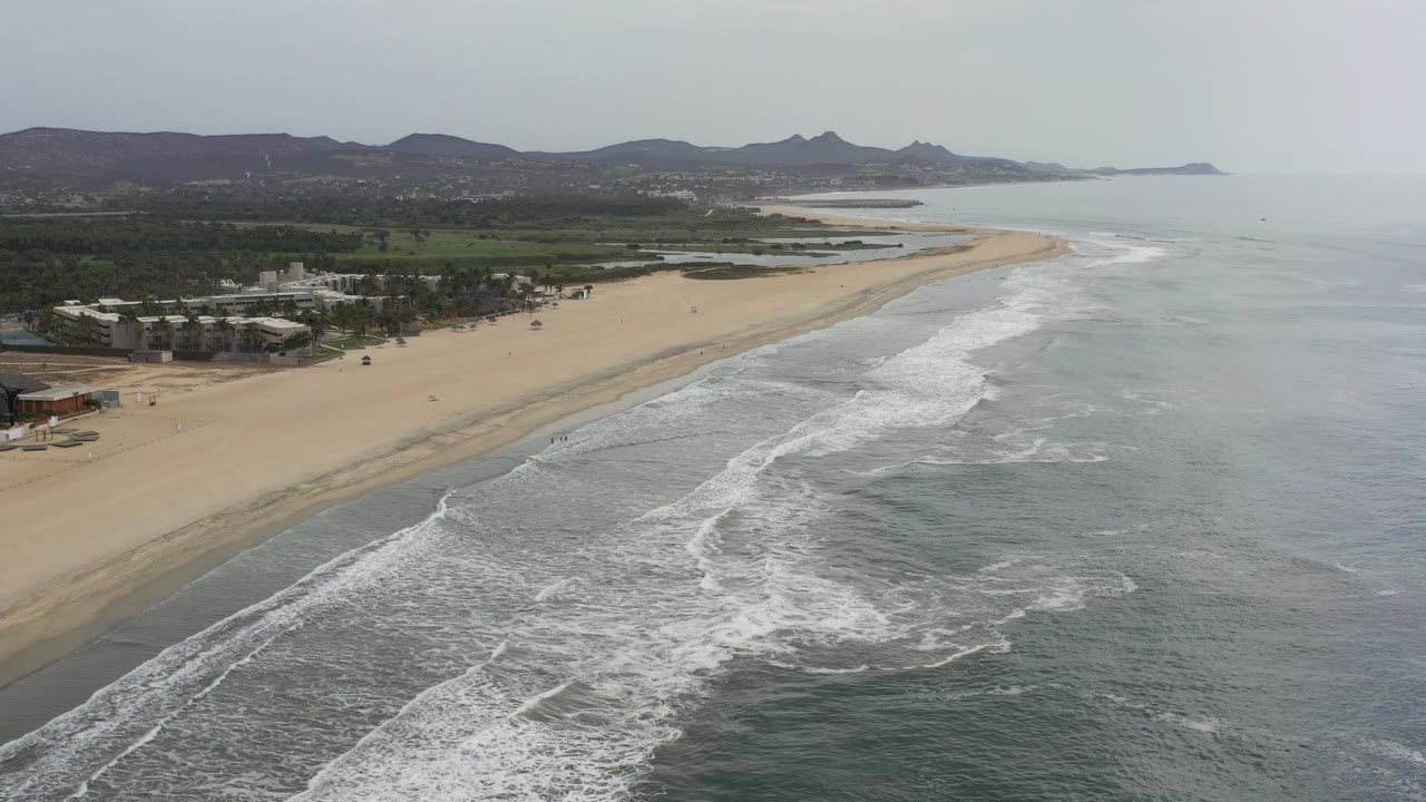 Aerial view over a Los Cabos beach with foamy surf in the foreground, sand and low buildings to the left, distant hills under a hazy sky, and no clearly visible people.