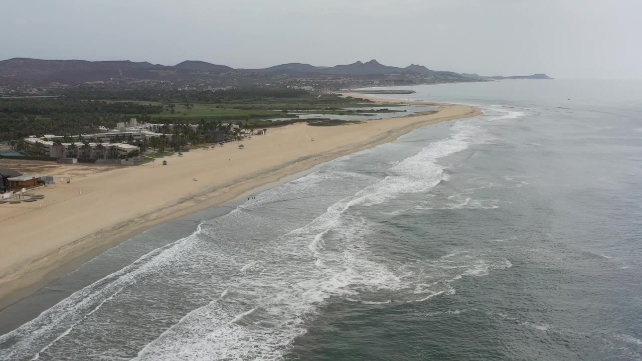 Wide aerial frame of the Los Cabos shoreline with white breakers crossing gray-green water, a broad sand strip, clustered buildings at left, dark hills, and no clearly visible people.