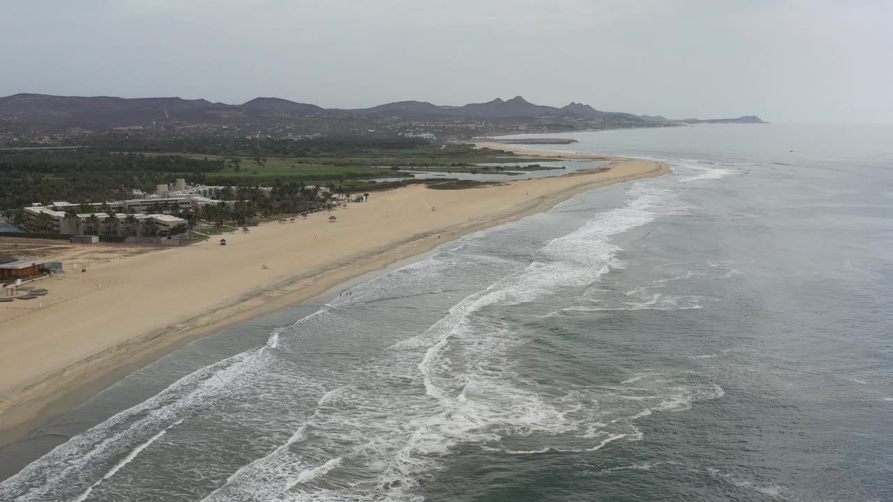 Coastline aerial with foamy water, sand, greenery, and mountains.