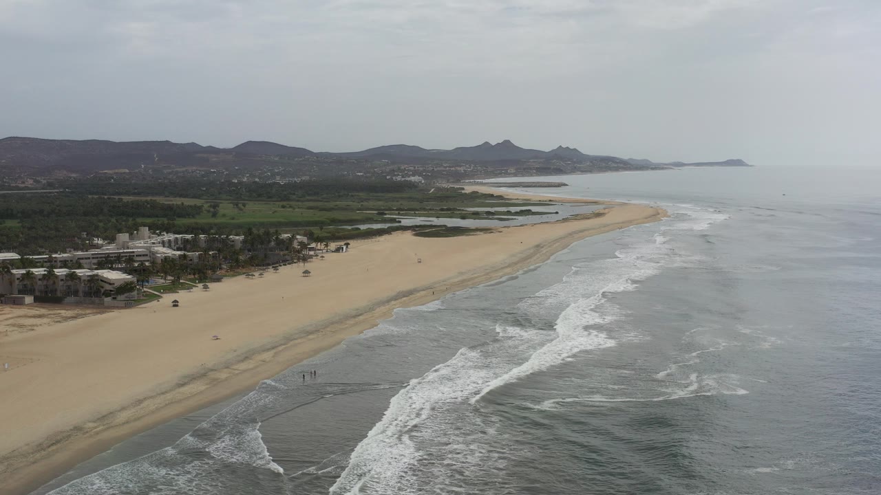 Aerial view of waves reaching a wide sandy beach with coastal buildings and hills beyond.