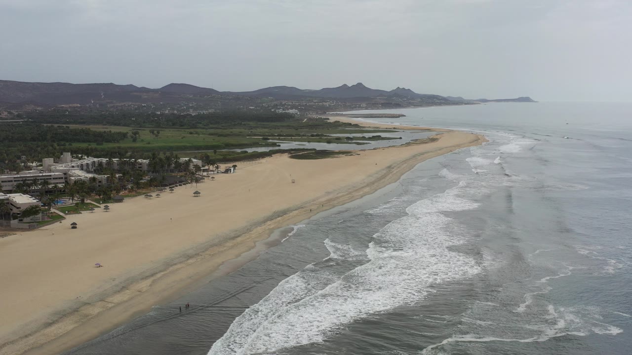 Wide aerial frame of a Los Cabos, Mexico sandy beach with white surf lines in the foreground, green coastal land to the left, distant mountains, and no visible people.
