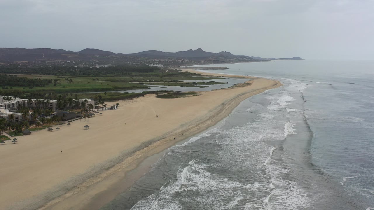 Aerial view over a Los Cabos beach with tan sand curving along gray surf, green inland areas at left, distant hills on the horizon, and no visible people.