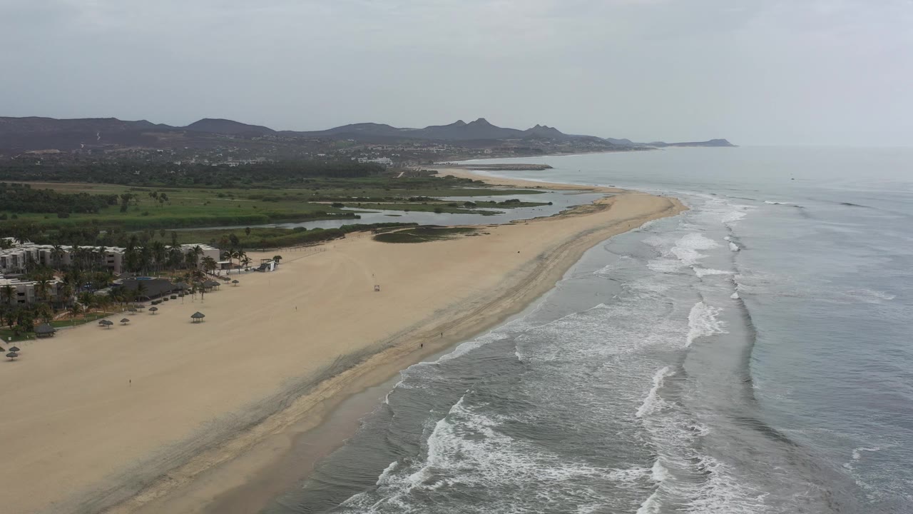 Wide aerial coastline with empty beach, surf, greenery, and mountains in haze.