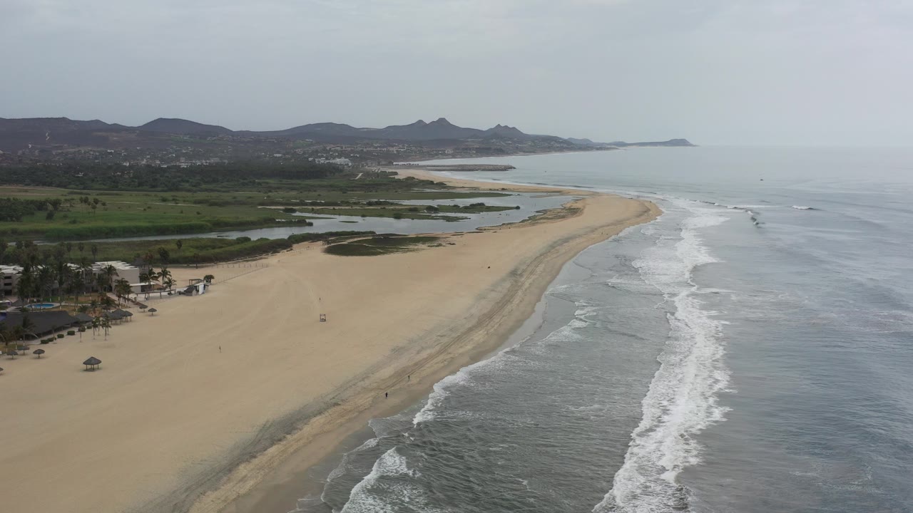 Opening aerial view over a Los Cabos beach with pale sand on the left, gray-blue surf on the right, green inland terrain beyond, distant hills, and no visible people.