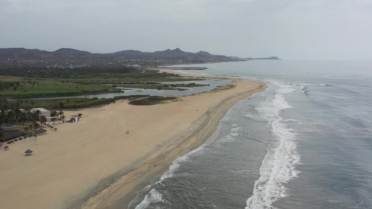 Curving beach with ocean waves and low hills under hazy daylight.
