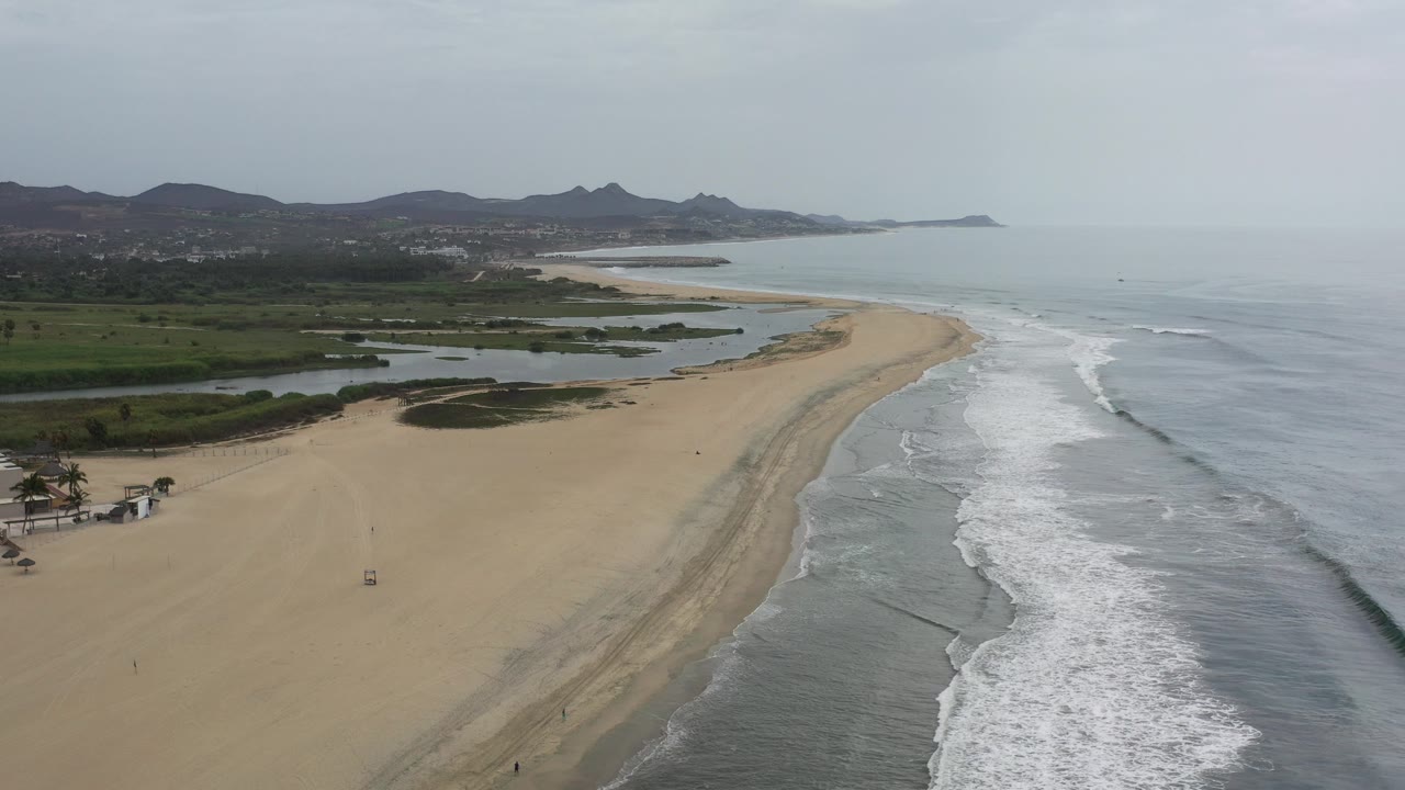 Wide aerial view of a Los Cabos beach with gray-blue surf on the right, pale sand below, inland lagoons to the left, distant hills, and no visible people.