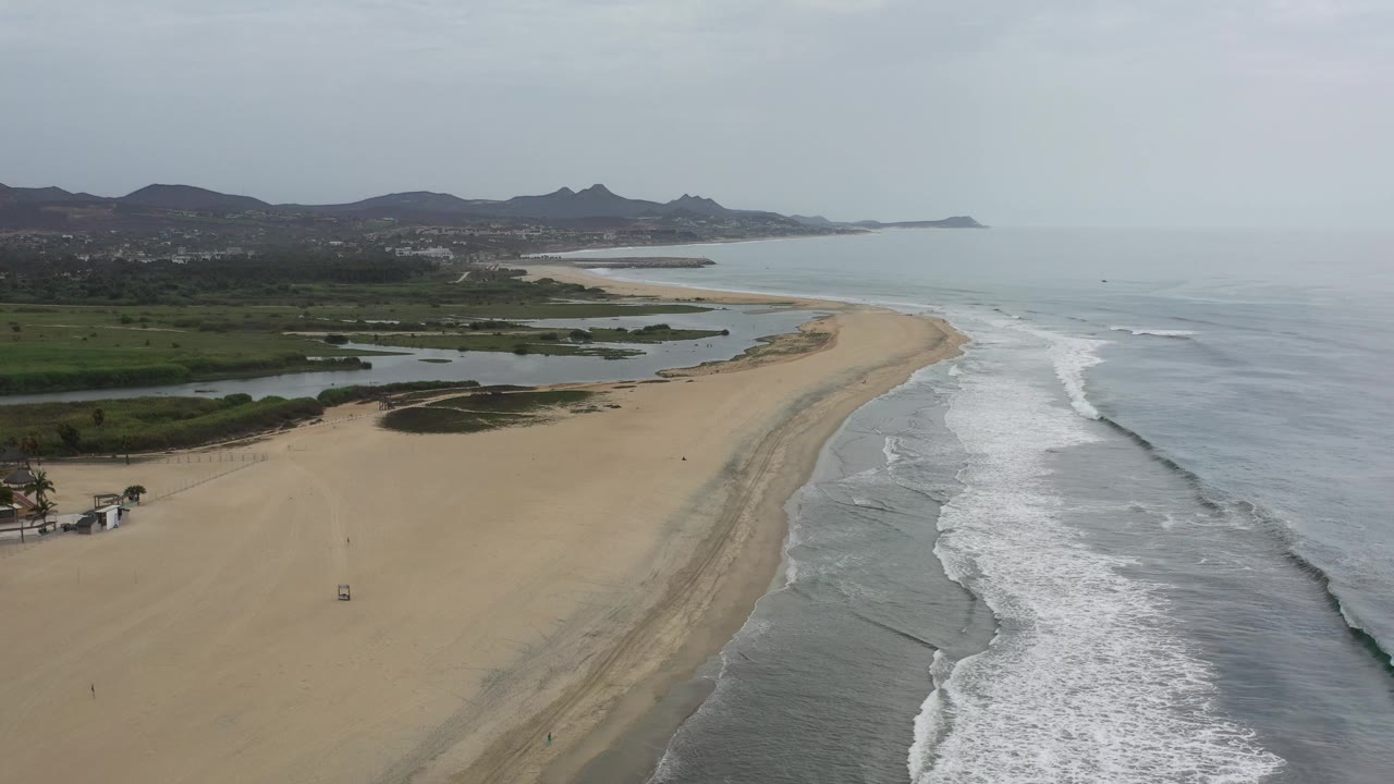 Curving beach with white waves, inland wetlands, and hazy hills.