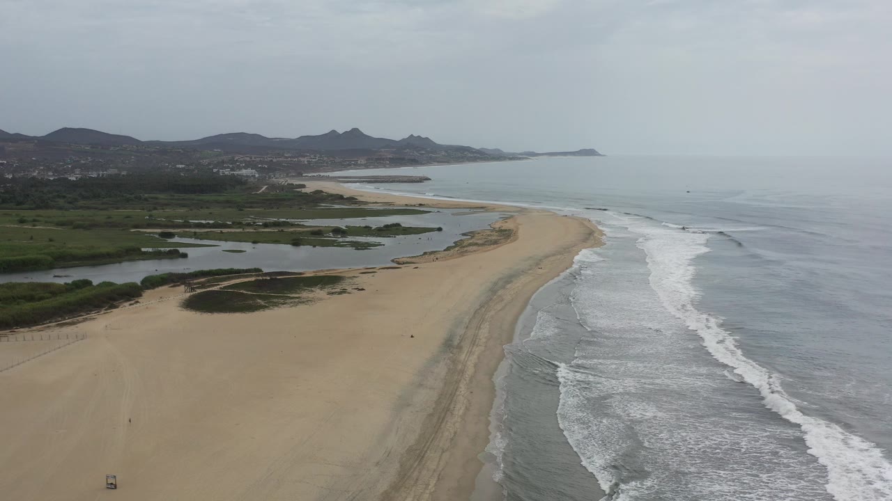 High aerial view over a curving sandy beach in Los Cabos, Mexico, with gray surf on the right, inland water and green vegetation on the left, distant hills, and no visible people.