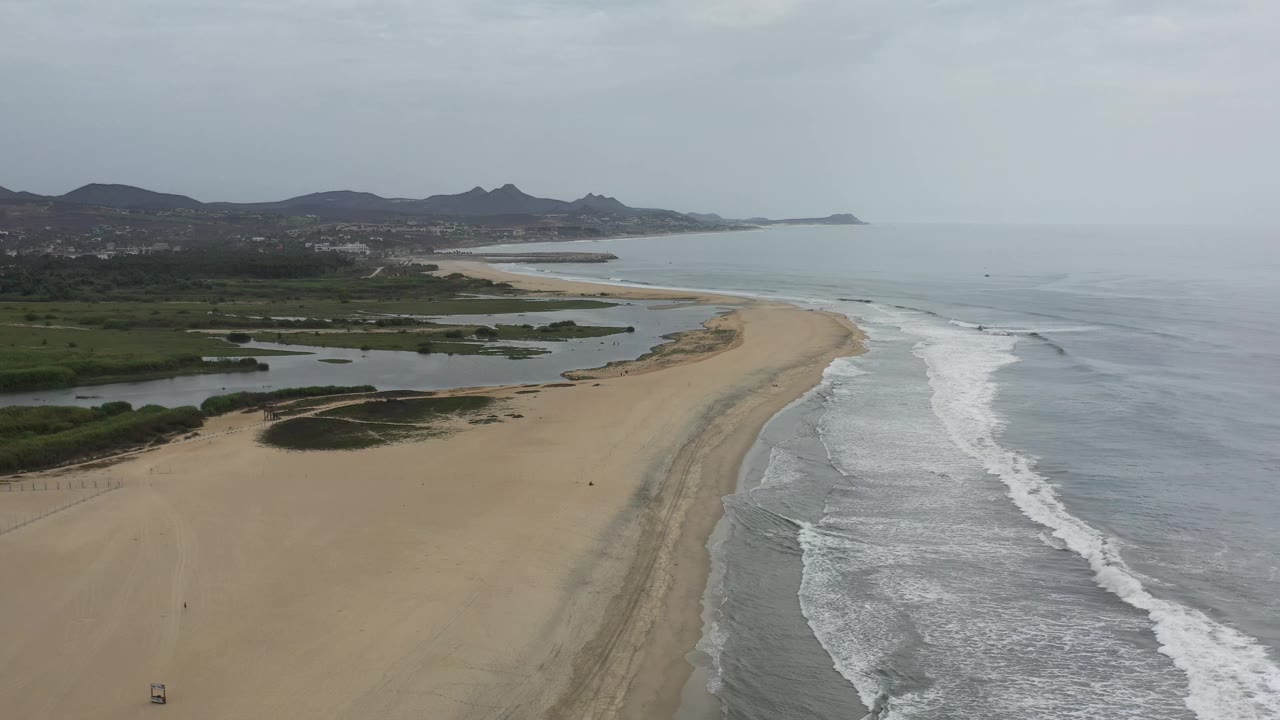 Aerial frame from Los Cabos, Mexico, showing the beach arc slightly farther along the shoreline, with white waves breaking parallel to shore and no visible people.