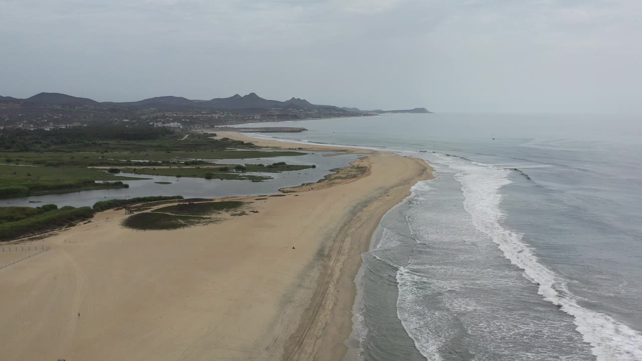 Curved shoreline with surf, sand, vegetation, and distant hills.