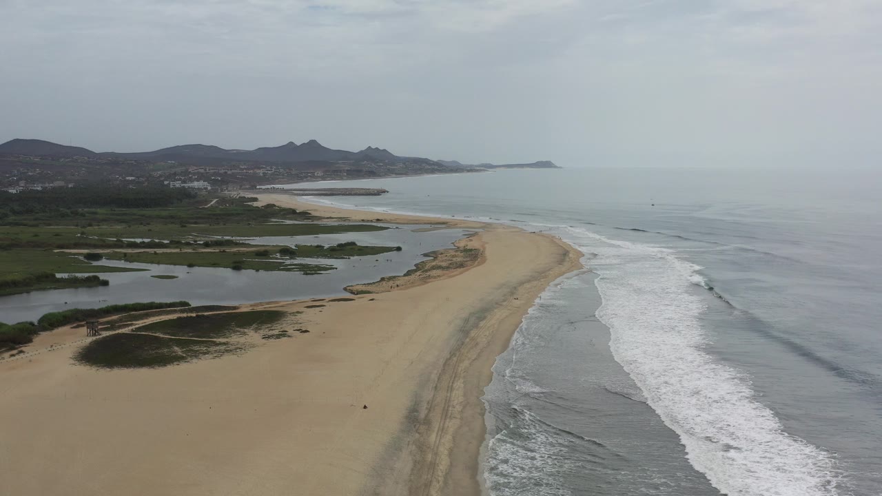 Aerial view over a Los Cabos beach where tan sand curves between lagoon water and gray ocean surf, with low mountains in the distance and no visible people.