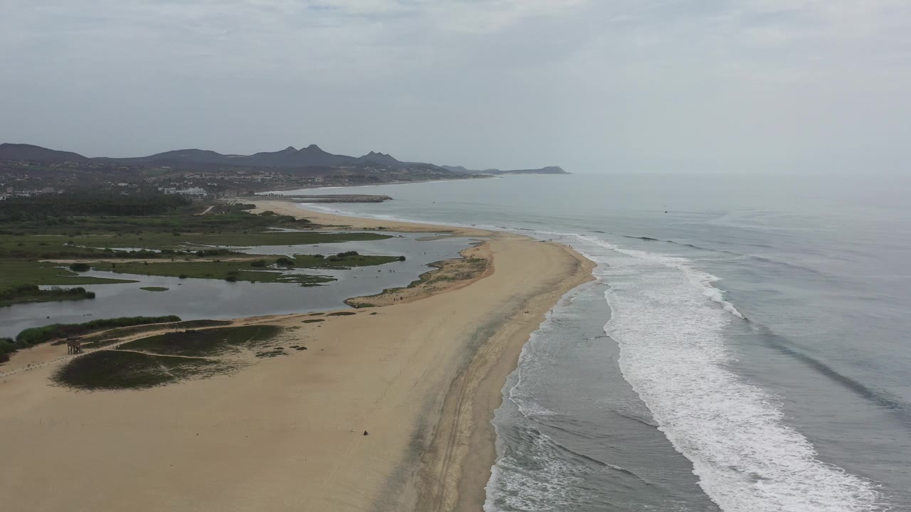 Drone-like view along an empty beach with surf, lagoons, and low mountains.