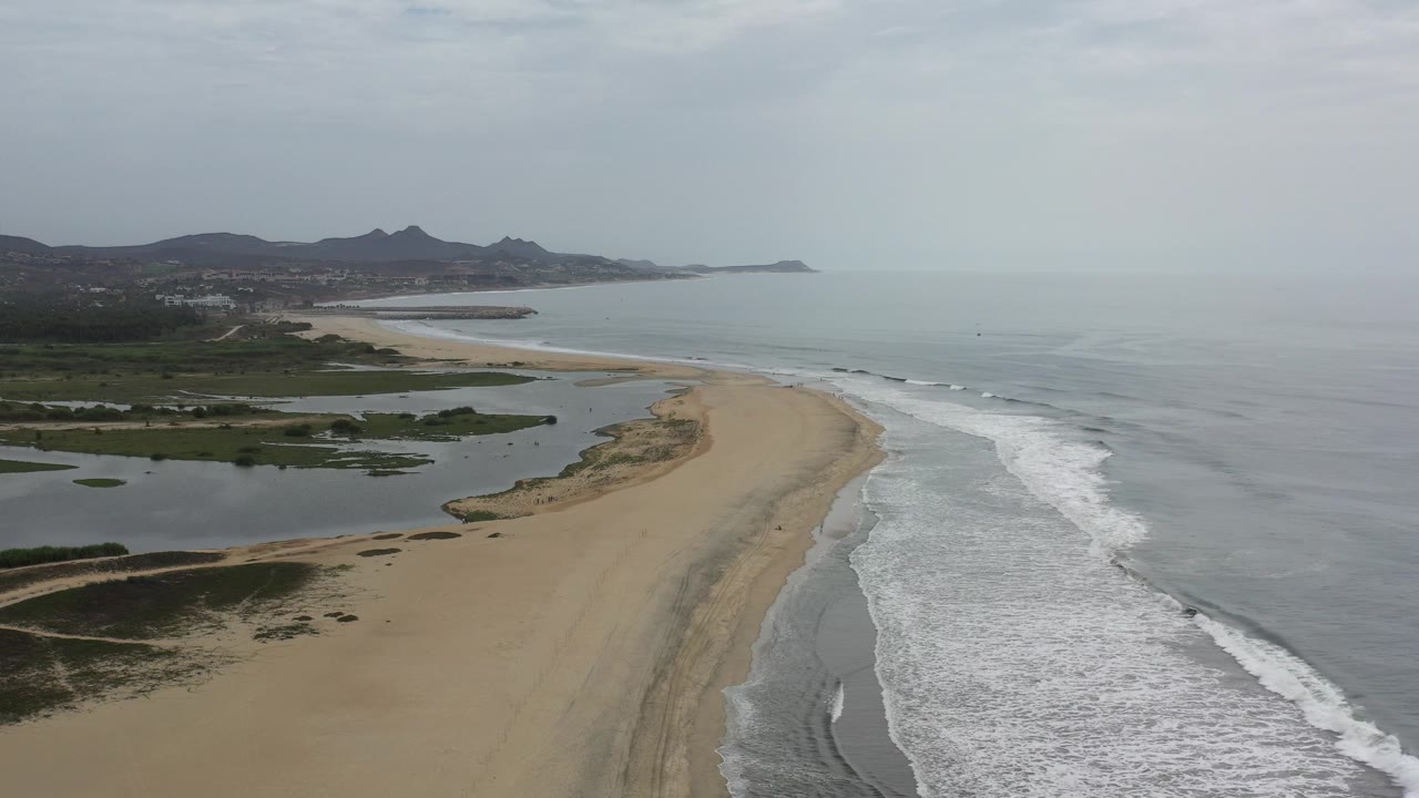 Opening aerial view over a Los Cabos beach with a wide sandbar, grey-blue surf on the right, lagoon water on the left, distant mountains, and no visible people.