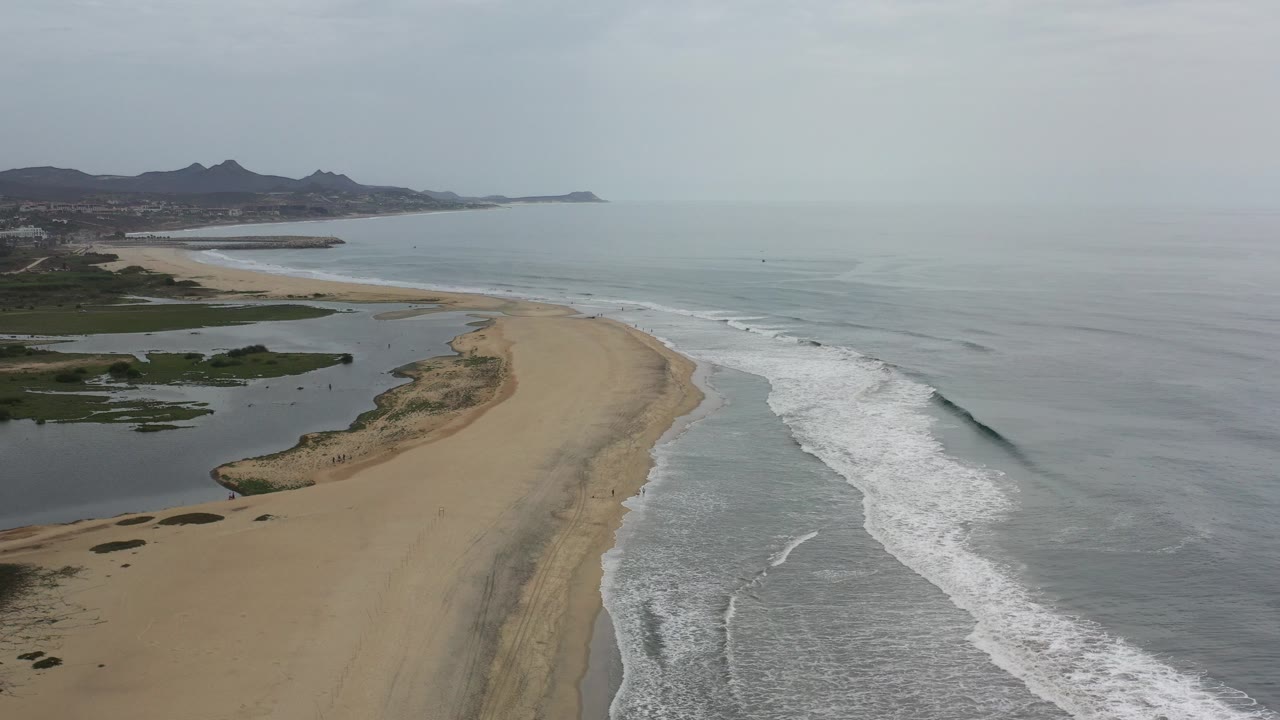 Opening aerial frame over a Los Cabos beach spit, with gray-blue surf on the right, darker lagoon water on the left, distant hills, and no visible people.