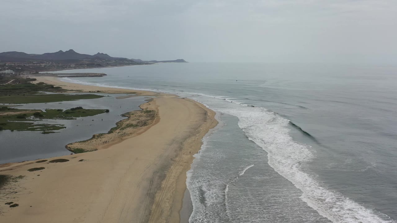 Aerial Los Cabos coastline view one second in, showing the surf band slightly farther along the sandbar, lagoon water to the left, and no visible people.