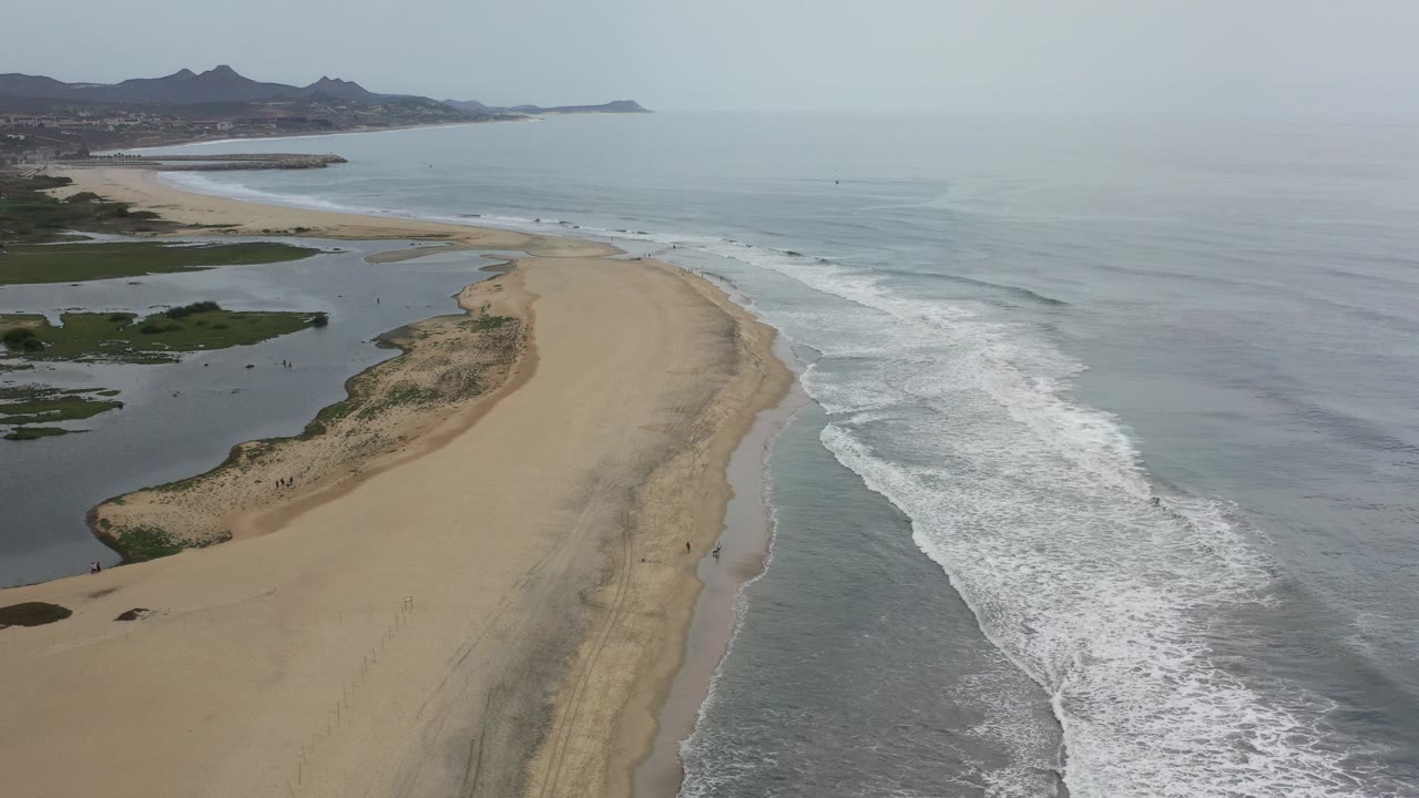 Aerial view over a Los Cabos sandy spit, with white surf on the right, darker lagoon water on the left, hazy mountains in the distance, and no visible people.