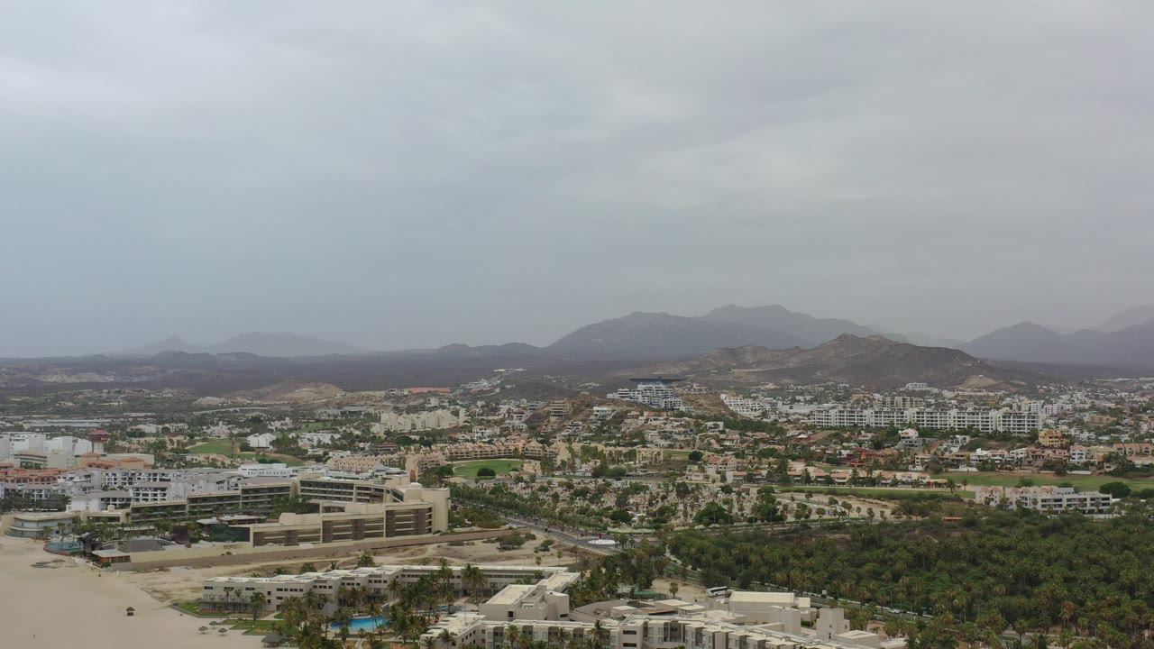 Opening aerial frame over Los Cabos, Mexico, showing low-rise buildings, green tree cover, roads, sandy ground, distant rugged hills, and no visible people.