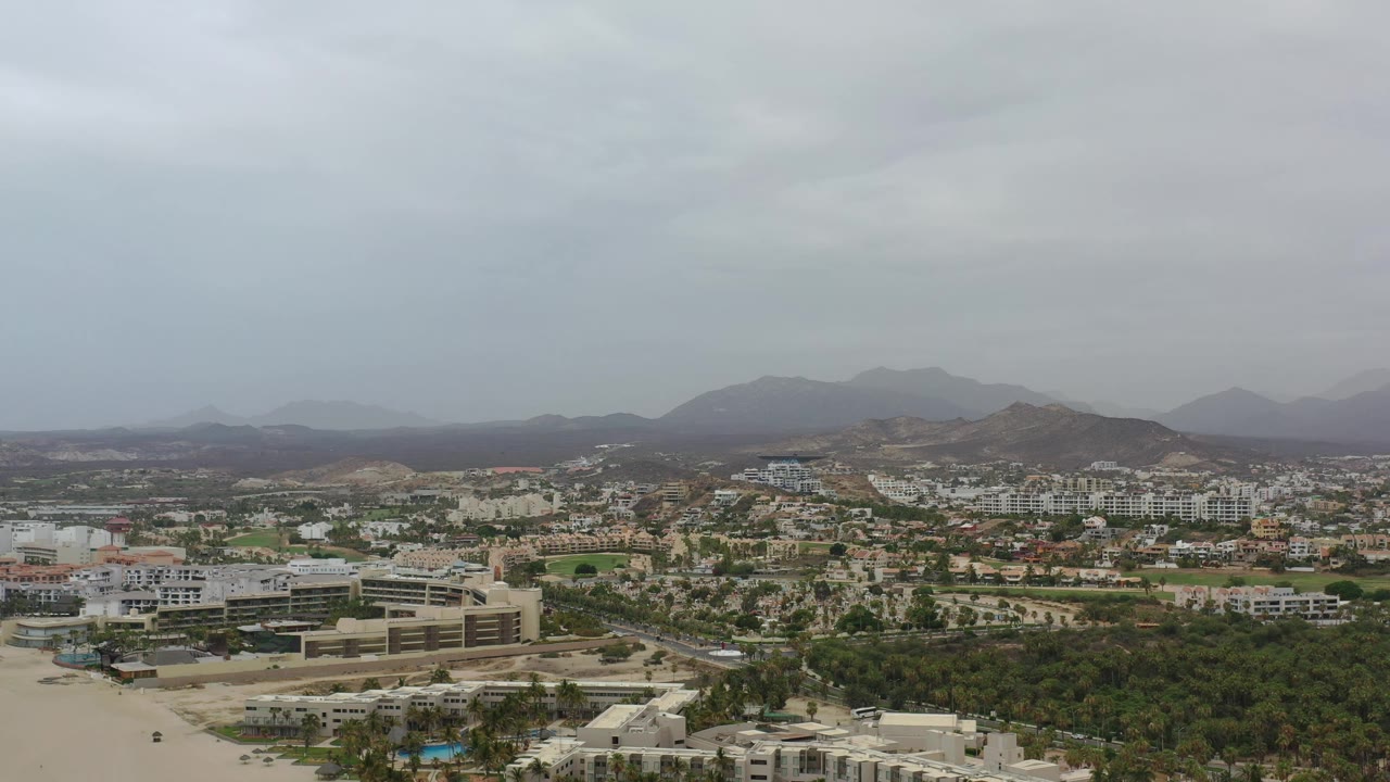 Early aerial view of Los Cabos, Mexico, with a broad tree-filled foreground, clustered buildings in the midground, hazy mountain ridges, and no visible people.