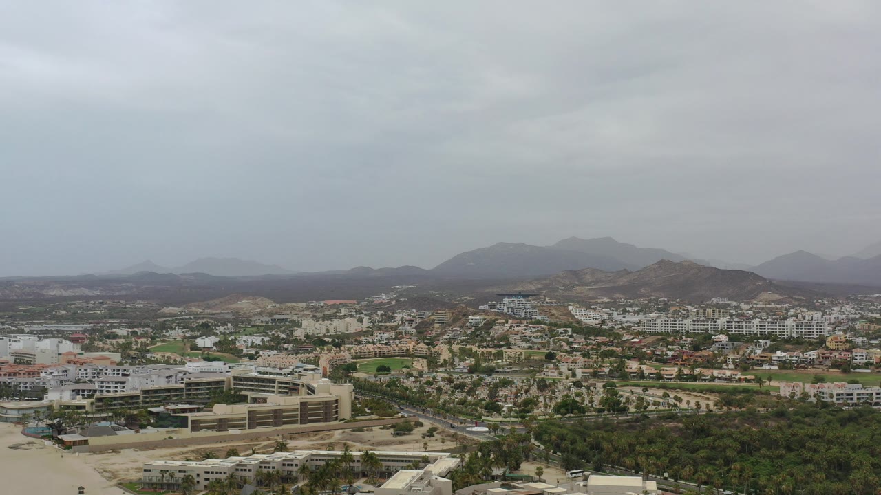 Opening aerial view over Los Cabos, Mexico, with low-rise buildings in the foreground, green tree cover, distant hills, gray overcast sky, and no visible people.