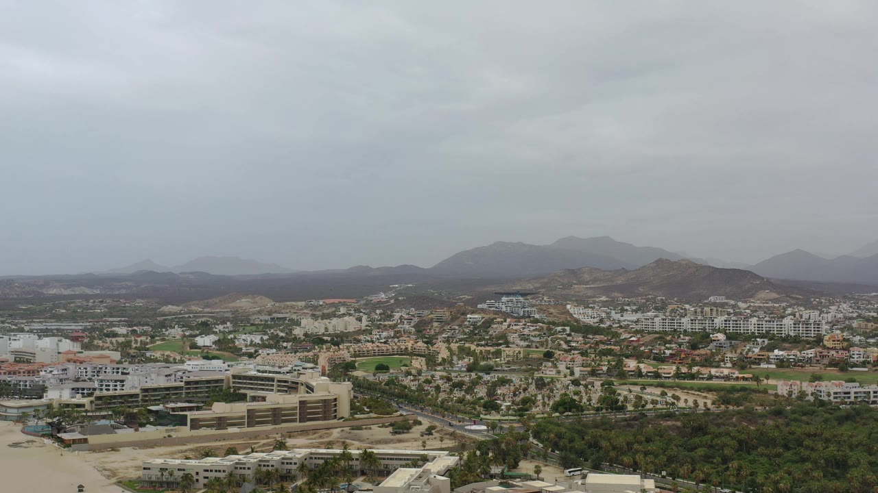 Aerial Los Cabos view one second in, showing a broad residential area, clusters of white buildings, tree-lined ground, low hills, cloudy light, and no visible people.