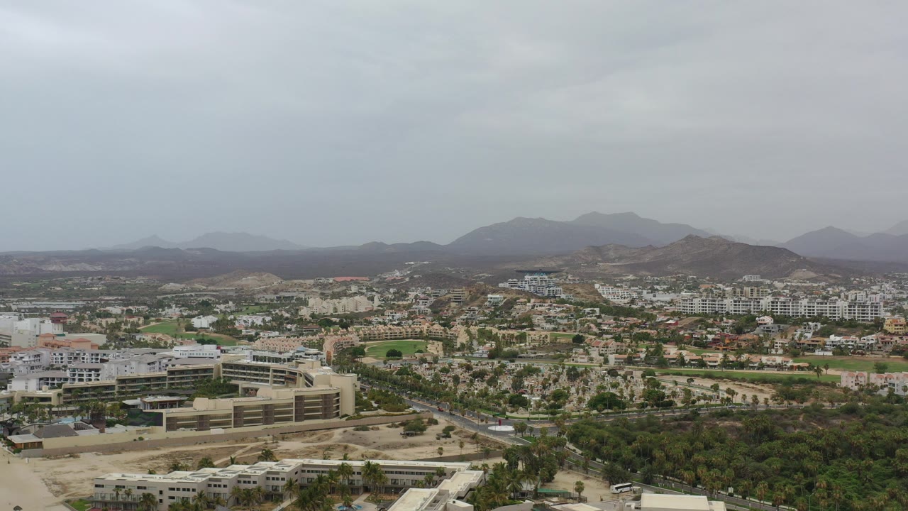 Opening aerial frame over Los Cabos, Mexico, showing foreground rooftops, landscaped palms, low-rise development, distant desert mountains, and no visible people.