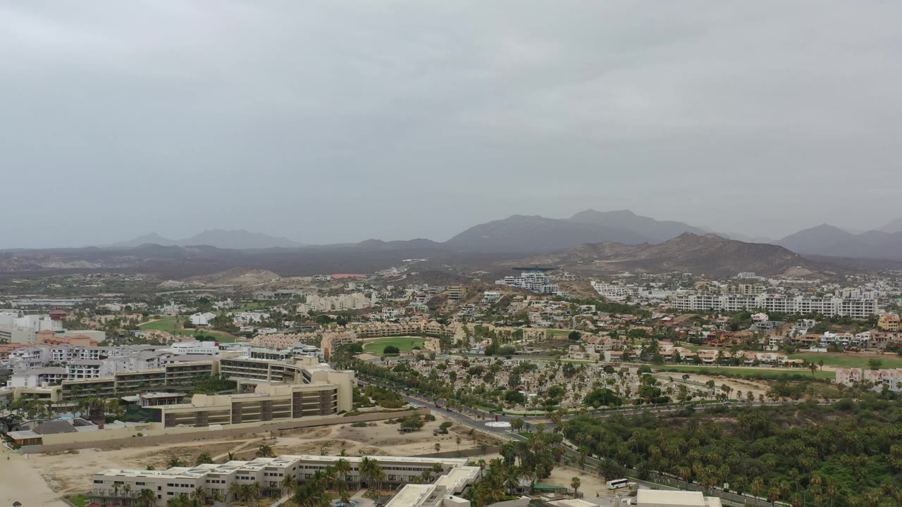 Aerial frame over Los Cabos with a broad band of gray sky above resort-style buildings, green landscaping, dry terrain, and no visible people.