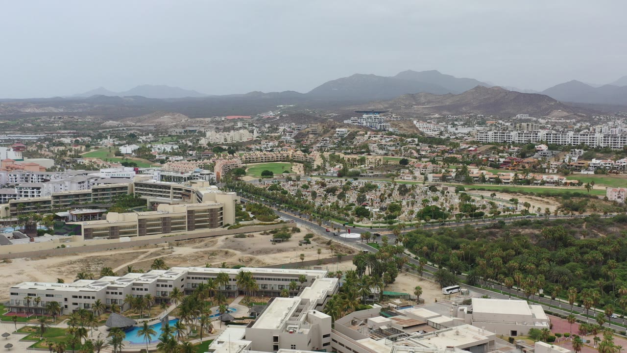 Aerial view over low-rise buildings, palm trees, roads, and distant mountains.
