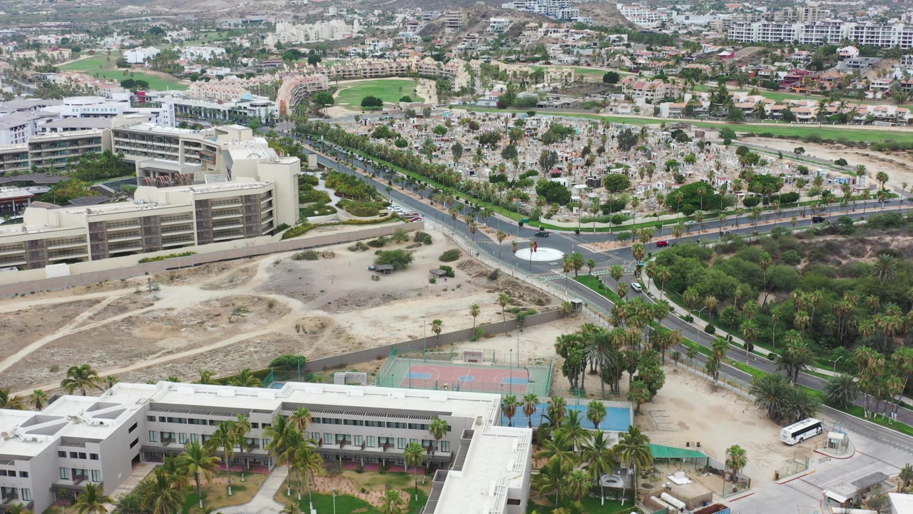 High oblique aerial view in Los Cabos, Mexico, with a divided road, light-colored buildings, tennis courts, palm trees, dry open ground, and no visible people.