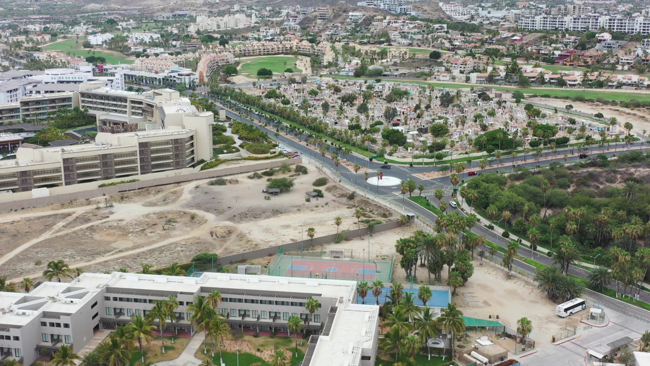Aerial frame in Los Cabos, Mexico, showing cars along the road, a large modern building, colorful tennis courts, palms, sandy open lots, and no visible people.