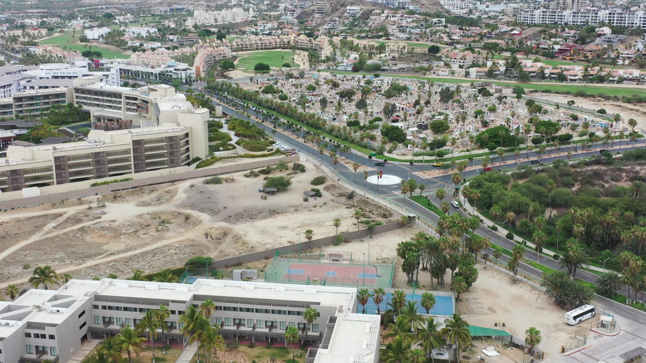 Drone-height view over a roundabout, roadway, buildings, palms, and courts.