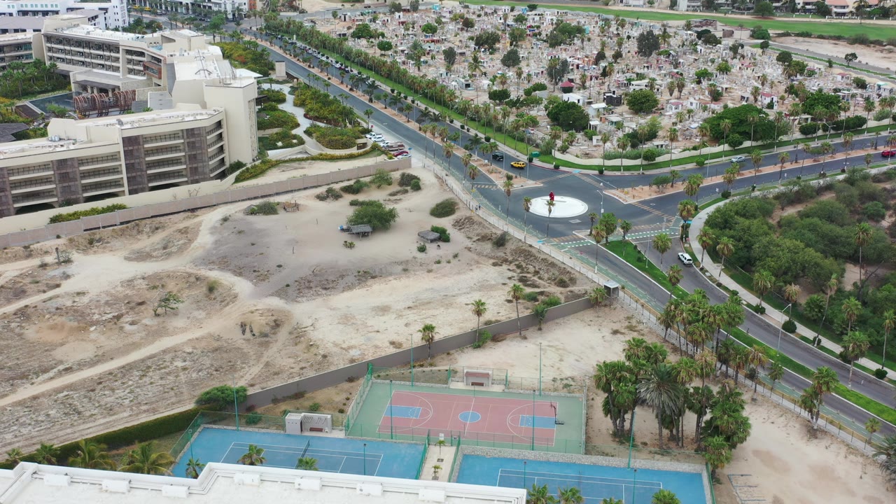 Aerial frame in Los Cabos, Mexico, with the circular traffic island closer to center, sports courts along the lower edge, a large building to the left, dry open lots, and no visible people.