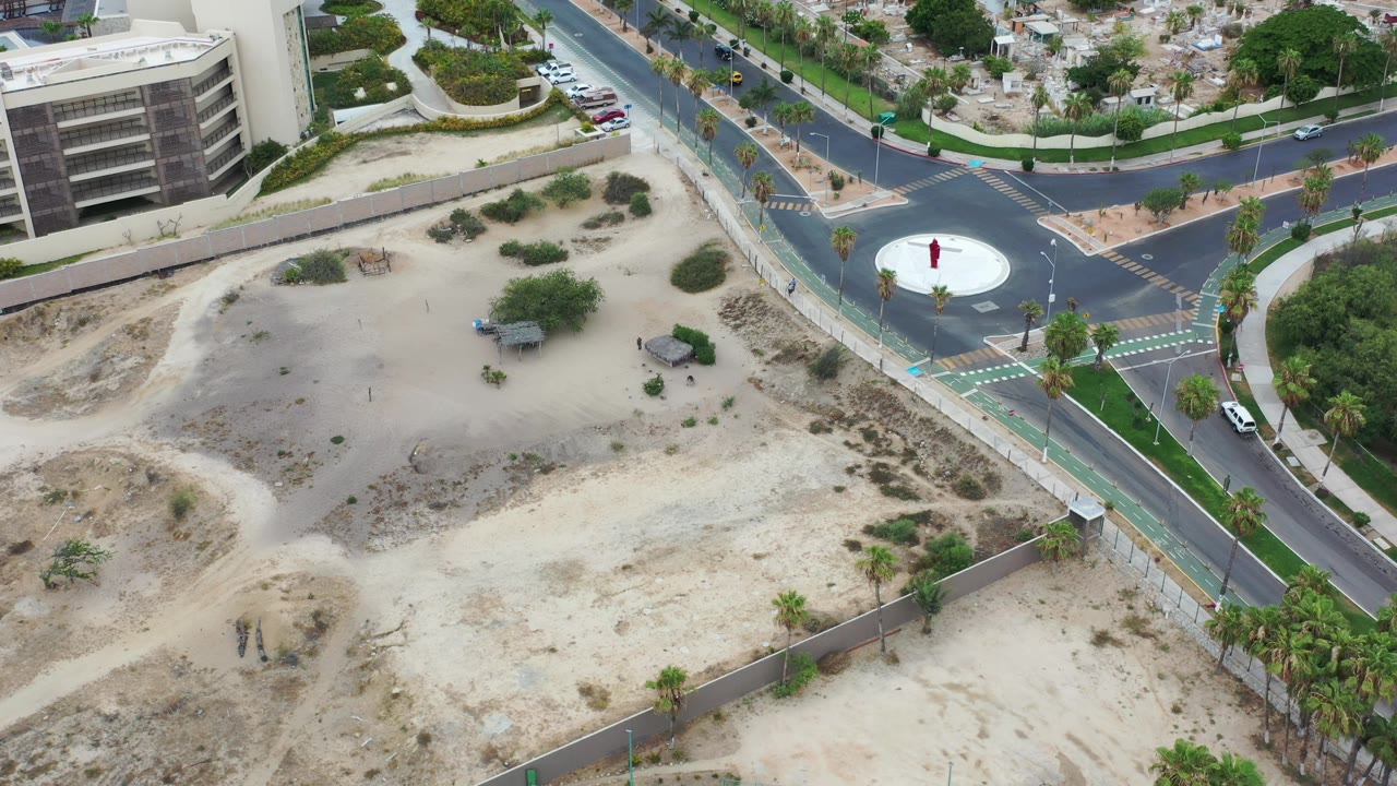 Los Cabos aerial frame with the roundabout near the upper right, a large sandy lot below it, palm-lined roads, modern buildings at the edge, and tiny visible people.